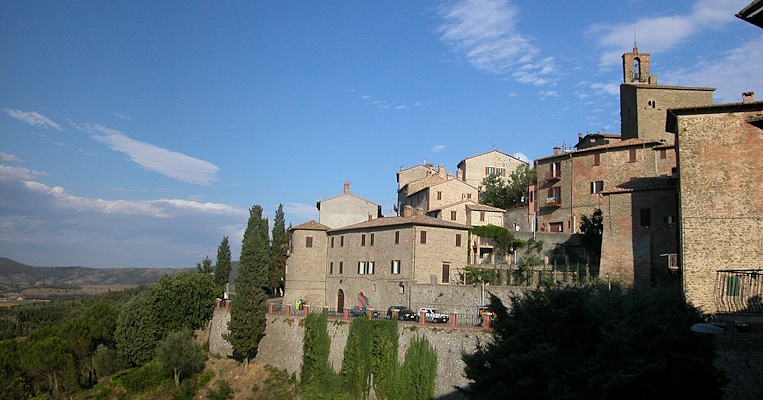  Medieval walls stand on a verdant slope, surrounded by stone houses and cypress trees, with a vast panorama in the background. 