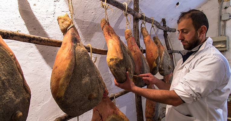 Jambons de Norcia IGP suspendu pour maturation avec un charcutier qui vérifie la maturation du jambon à l’aide d’un bâton.