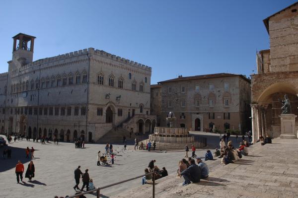 Piazza IV Novembre with its major fountain and Palazzo dei Priori, passers-by enjoy the sunny day sitting on the steps of the cathedral or strolling along.