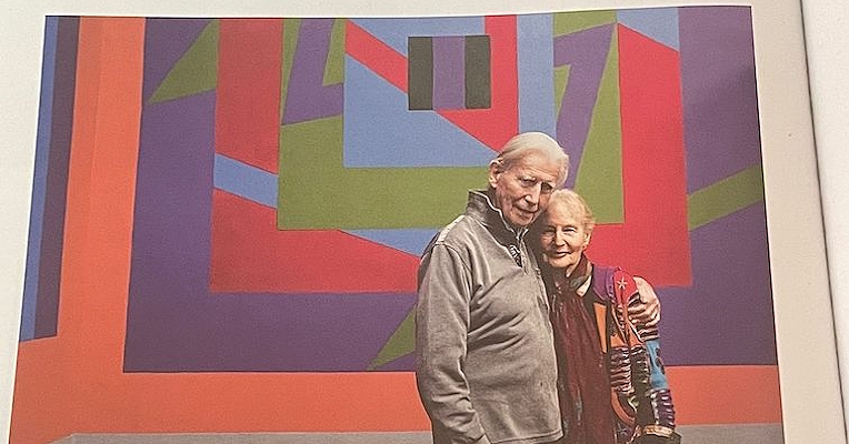 An elderly couple embracing in front of a wall decorated with colorful geometric shapes.