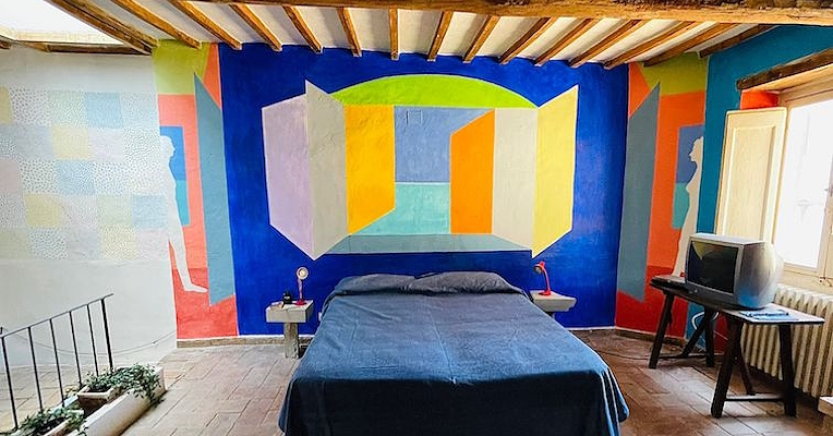 Bedroom with a blue wall decorated with colorful geometric shapes behind the bed and exposed ceiling beams.