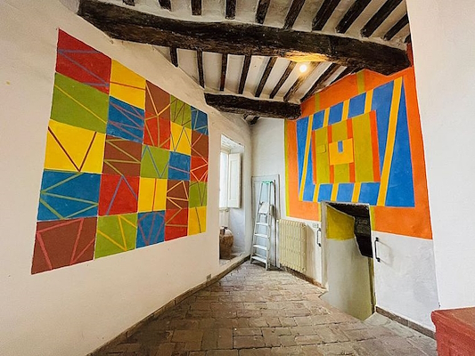 Hallway with exposed beams and walls decorated with colorful geometric patterns, a ladder, and a window.