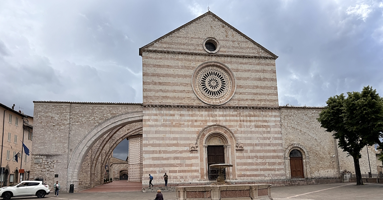 Fassade der Basilika der Heiligen Klara in Assisi, mit Rosette und seitlichem Bogen, Blick vom Platz mit Brunnen im Vordergrund.