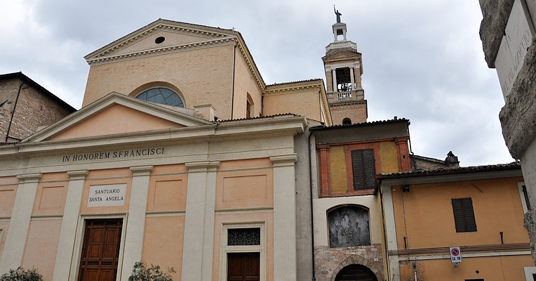 Fassade der Kirche des Heiligen Franziskus in Foligno mit Glockenturm im Hintergrund, teilweise Sicht auf angrenzende Gebäude.