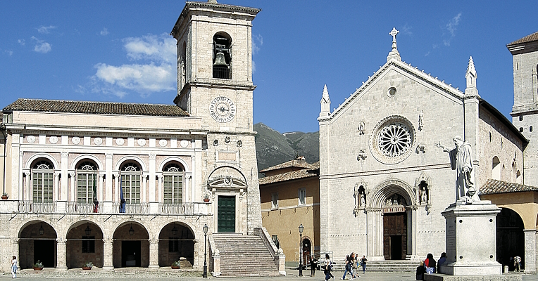 Zentraler Platz mit dem Rathaus und der Basilika des Heiligen Benedikt in Norcia, mit Glockenturm und Statue im Vordergrund.