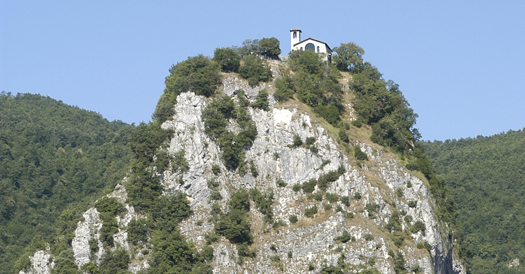 Felsvorsprung mit Vegetation und kleiner Kirche auf der Spitze, eingebettet in eine grüne Hügellandschaft unter klarem Himmel.