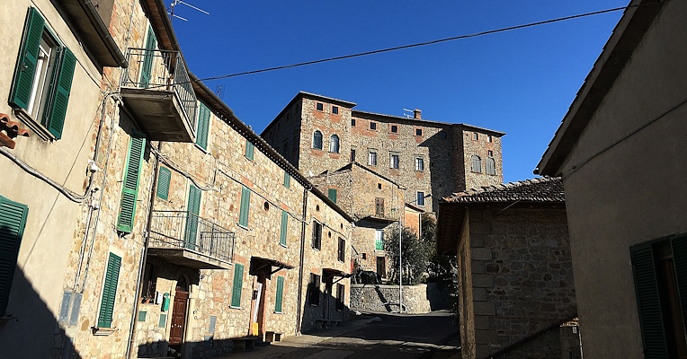 Straße im Dorf Carnaiola mit Steinhäusern und grünen Fensterläden, unter klarem Himmel; Burg am Ende der Straße.