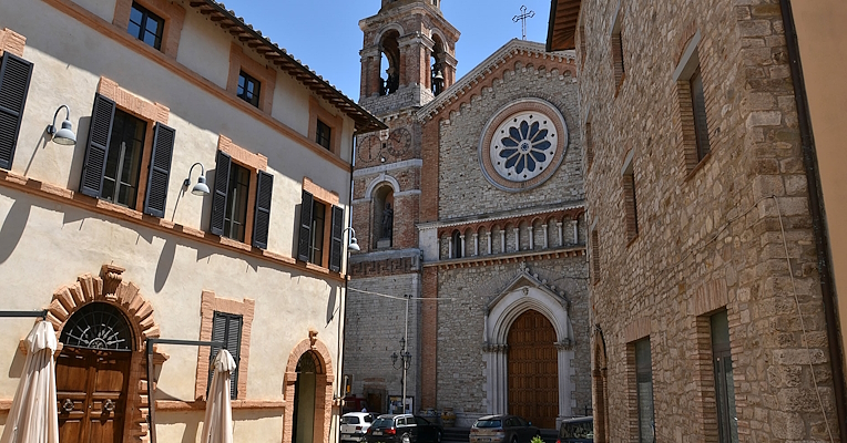 View of a square with stone and brick buildings, dark-shuttered windows, and a church with a rose window and bell tower in the background.