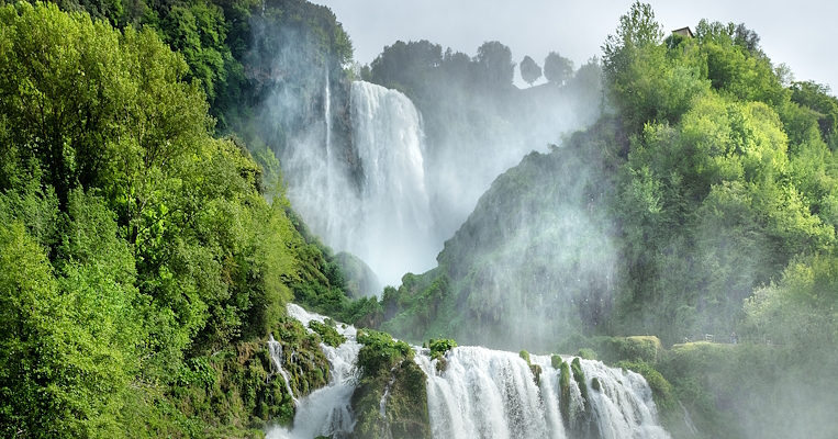  Cascata circondata da rigogliosa vegetazione verde, con giochi d'acqua e vapori, sotto un cielo limpido e azzurro. 