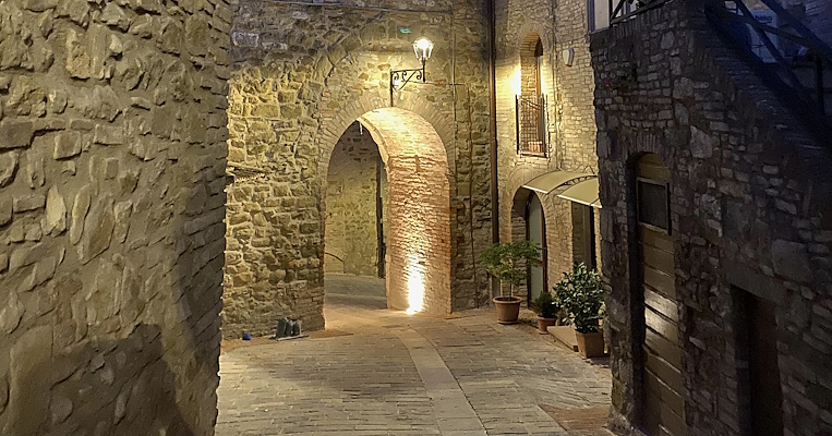  Vicolo in pietra illuminato da una luce calda, con un arco suggestivo e un campanile sullo sfondo al crepuscolo. 