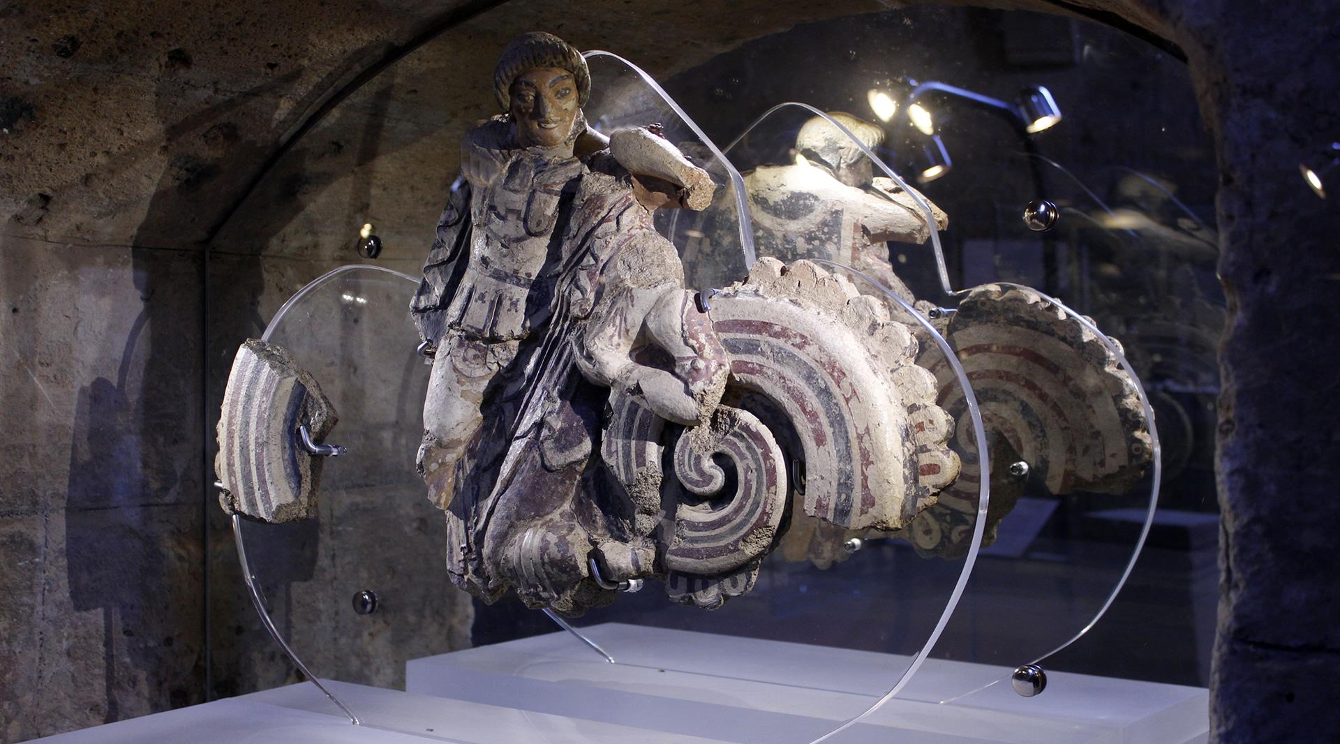 Restes d'une sculpture polychrome en terre cuite représentant une figure humaine avec des détails décoratifs complexes. La sculpture est exposée dans une vitrine illuminée, mettant en valeur les couleurs vives et les décorations complexes de l'objet.